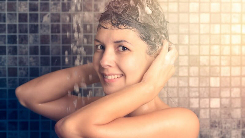 Frau steht unter der Dusche und massiert Shampoo in die Haare ein, blickt in die Kamera