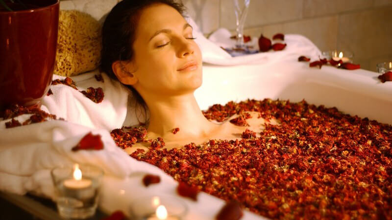 Frau mit geschlossenen Augen in Badewanne mit Kerzen und Sektglas beim Rosenblütenbad mit Rosenblättern