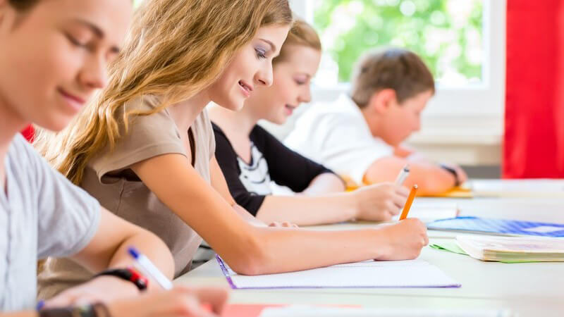 Vier Schüler/innen sitzen in einer Reihe im Klassenzimmer und schreiben eine Klassenarbeit