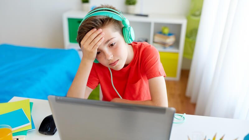 Junge in rotem Shirt sitzt mit grünem Kopfhörer vor dem Laptop im Kinderzimmer