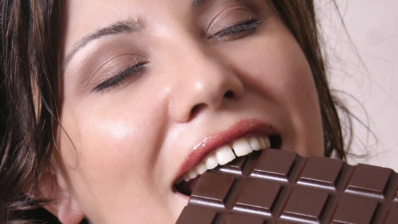 Dunkelhaarige Frau beisst genüsslich mit geschlossenen Augen in Tafel Schokolade