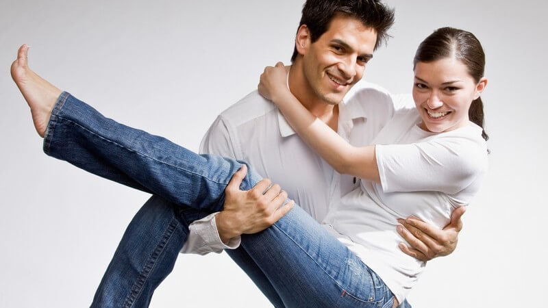 Ansicht von oben: Junger Mann trägt seine Freundin im Arm, beide barfuss, lächelnd