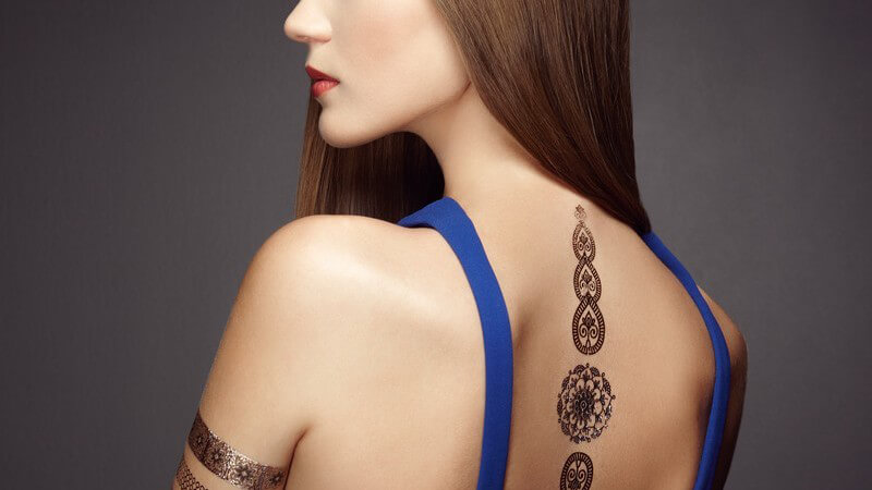 Rückansicht eines Models vor grauem Hintergrund, lange glatte und braune Haare, Tattoos auf Wirbelsäule und Oberarm
