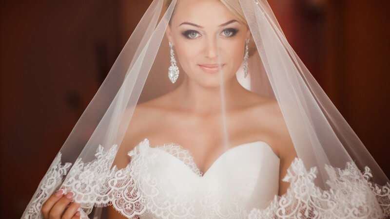 Hübsche blonde Braut mit langen glamourösen Ohrringen unter einem durchsichtigen Schleier