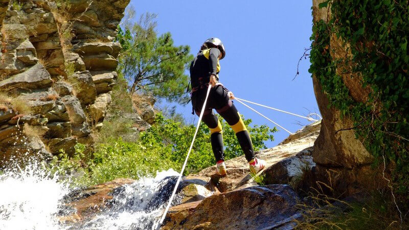 Frau klettert gesichert zwischen Felsen mit kleinem Wasserfall, in schwarz-gelber Schutzkleidung mit Helm