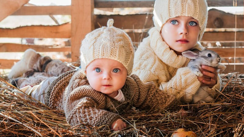 Kleines Mädchen mit Kaninchen auf dem Arm und ein Baby sitzen in einem Stall im Heu, warme Wollmützen an