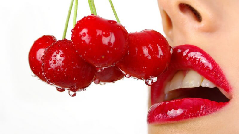 Nahaufnahme Frau mit rotem glänzendem Lippenstift hält rote frisch gewaschene Kirschen an ihren Mund