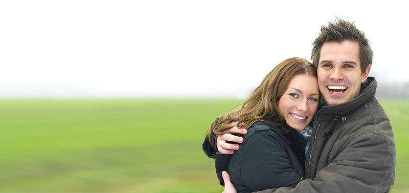 Junges Paar in Jacken umarmen sich, lächeln in die Kamera, hinten grüne Wiese