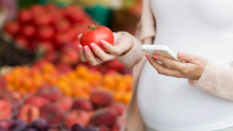 Schwangere Frau hält eine Tomate an einem Marktstand und guckt in ihr Smartphone