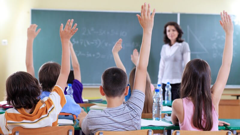 Rückansicht, Schüler in Klasse zeigen auf, vorne Lehrerin an Tafel