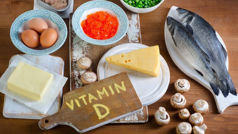 Vitamin D-haltige Nahrungsmittel wie Dorsch, Champignons, Butter, Käse, Erbsen und Eier, mit Beschriftung auf Holzbrett