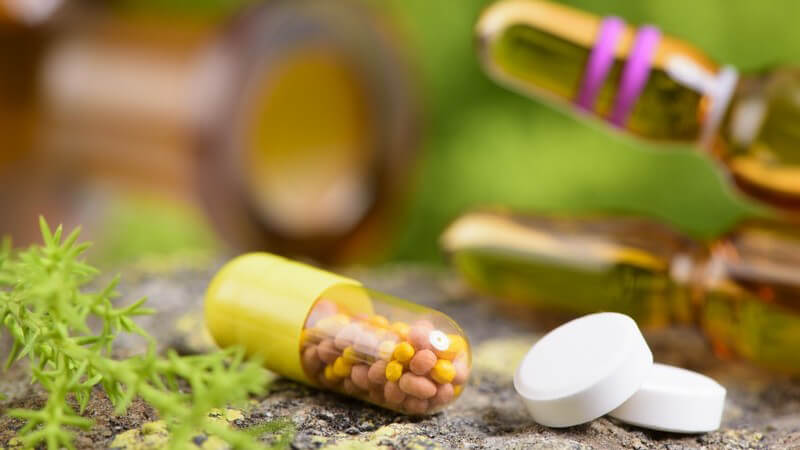 Zwei weiße Tabletten und eine gelbe Kapsel auf Stein, im Hintergrund liegen Ampullen