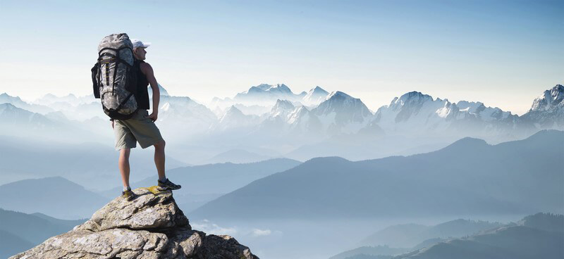 Tourist mit großem Wanderrucksack steht auf einem Berggipfel und schaut in die Berge