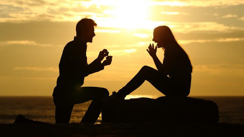 Silhouette eines Pärchens am Strand im Sonnenuntergang, er macht ihr einen Heiratsantrag