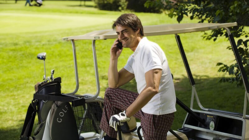 Golfspieler am Golfplatz neben seinem Golfwagen telefoniert mit seinem Handy