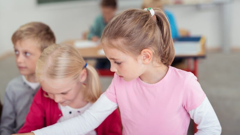 Mädchen in rosa Shirt auf weißem Longsleeve mit Mitschülern im Klassenzimmer