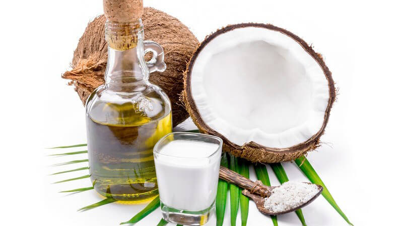 Kokosölflasche, Kokosmilchglas und Löffel mit Kokosraspeln neben Kokosnüssen vor weißem Hintergrund
