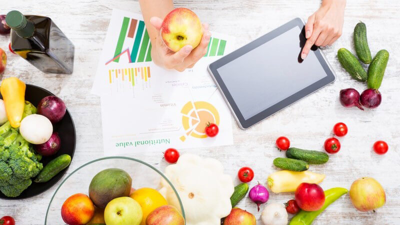 Tablett-PC auf Tisch mit buntem Obst und Gemüse, eine Hand hält einen Apfel