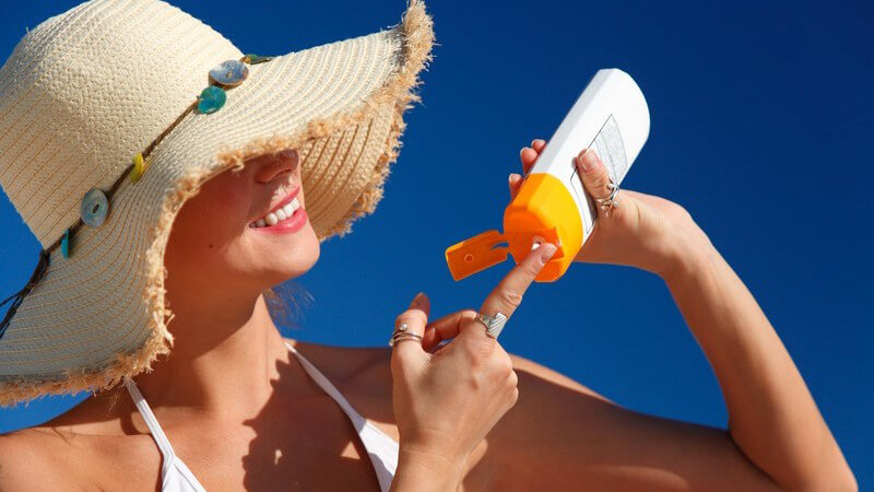 Frau mit Sonnenhut und weißem Bikini cremt sich mit Sonnencreme ein