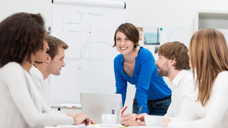 Junge Frau in blauem Oberteil steht an einem Whiteboard und spricht mit vier sitzenden Kollegen