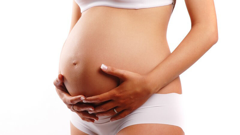 Schwangere Frau mit weißem BH und Slip hält sich den Bauch