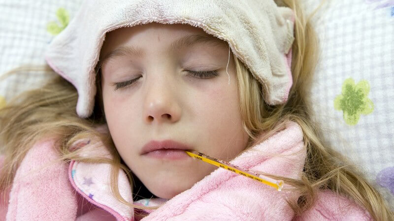 Warm eingepacktes kleines Mädchen liegt mit Fieberthermometer im Bett