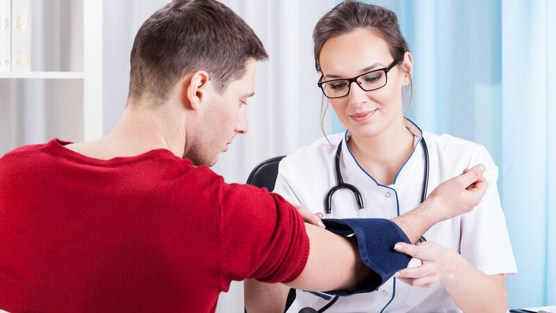 Junge Ärztin mit Brille misst den Blutdruck bei einem männlichen Patienten in rotem Pullover