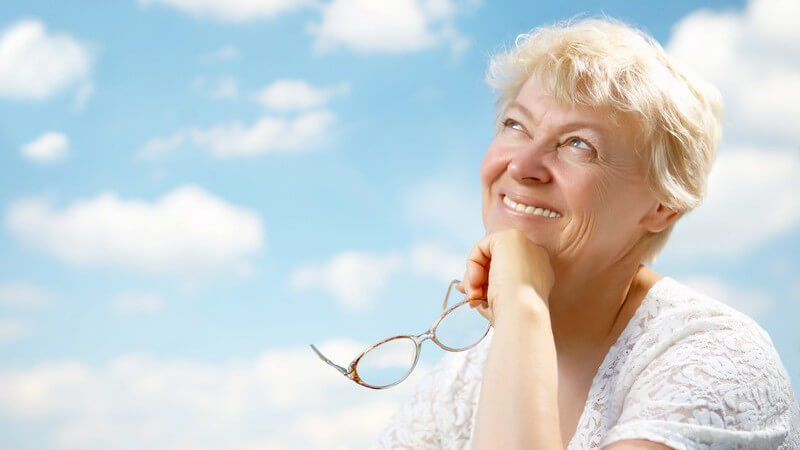 Ältere Frau mit Brille in Hand lächelt, schaut nach oben, blauer Himmel mit Wolken