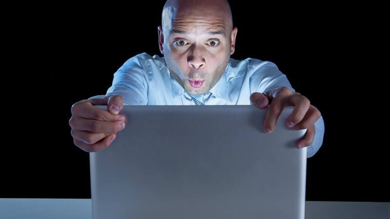 Kahlköpfiger Businessmann sitzt in dunklem Raum vor dem Laptop und hält ihn gespannt fest