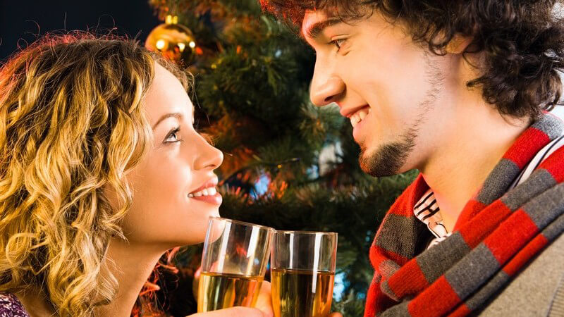 Junges Paar steht am Weihnachtsbaum, sie schauen sich lächelnd ins Gesicht und stoßen an