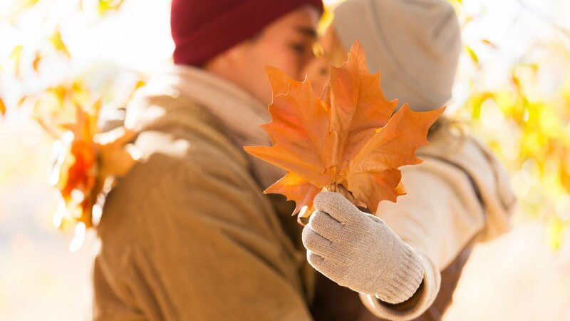 Küssendes Paar im Herbst, Frau verdeckt den Kuss mit Herbstblatt