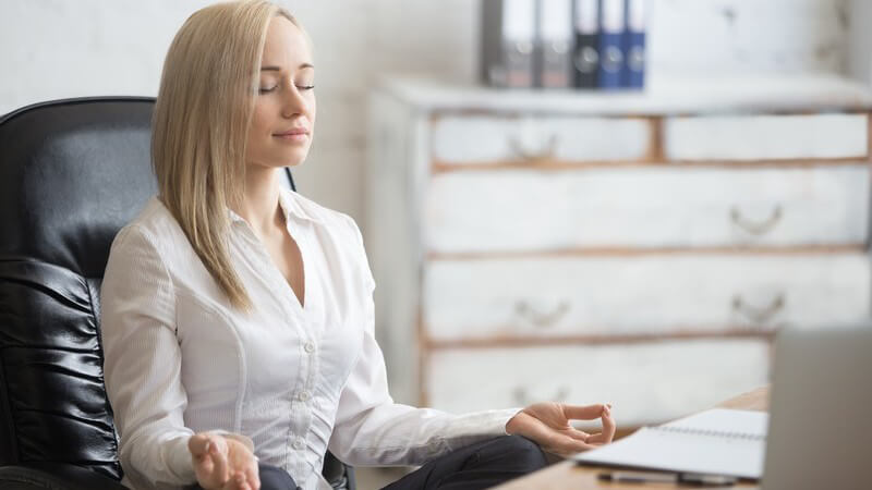 Blonde Frau in weißer Bluse meditiert oder macht Yoga am Schreibtisch im Büro