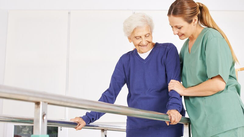 Frau in grünem Kittel hilft einer Seniorin in blauem Pullover auf Laufband