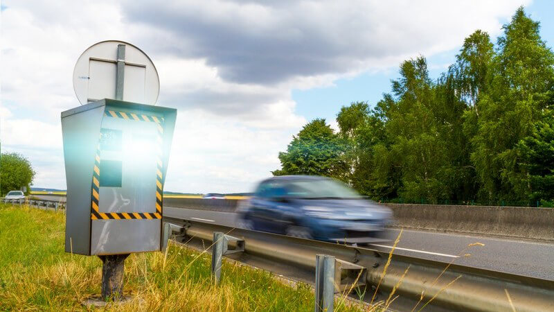 Stationäres Gerät zur Geschwindigkeitsüberwachung hinter der Leitplanke einer Schnellstraße, blaues Auto fährt vorbei