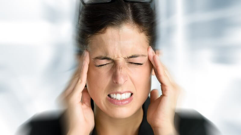 Frau vor verschwommenem Hintergrund fasst sich mit geschlossenen Augen und Kopfschmerzen an die Schläfen