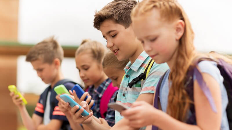 Fünf junge Schüler mit Schulrucksack befassen sich alle mit ihrem Smartphone
