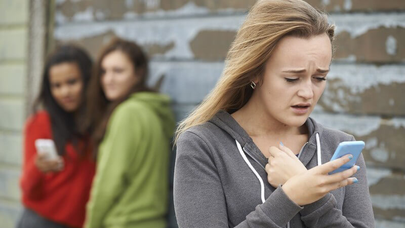 Jugendliches Mädchen liest eine Textnachricht auf ihrem Smartphone, im Hintergrund stehen zwei weitere Mädchen mit Handy