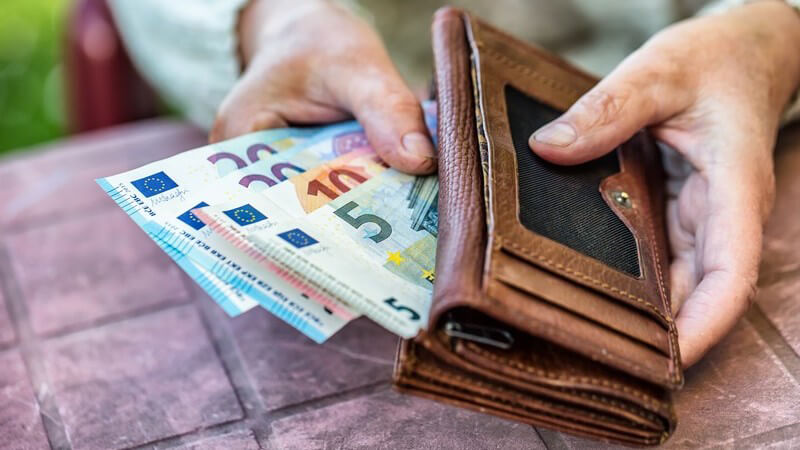 Seniorin sitzt an einem Tisch und hält eine braune Brieftasche mit Euroscheinen