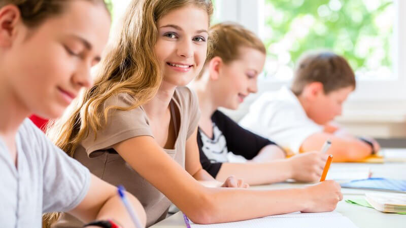 Vier Schüler beim Schreiben einer Klassenarbeit, Mädchen lächelt in die Kamera