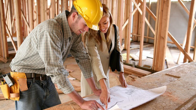 Handwerker o. Architekt mit Bauhelm und Werkzeug steht unter Holzgerüst mit Bauplan, junge Frau zeigt darauf