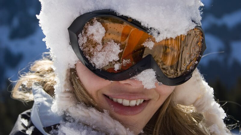 Blonde Frau mit verschneiter Kapuze und Skibrille lächelt in Kamera