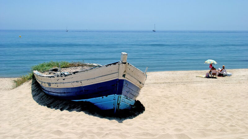 Altes blaues Boot am Strand von Andalusien, verwittertes Boot, mit Meer im Hintergrund und Strandgästen daneben