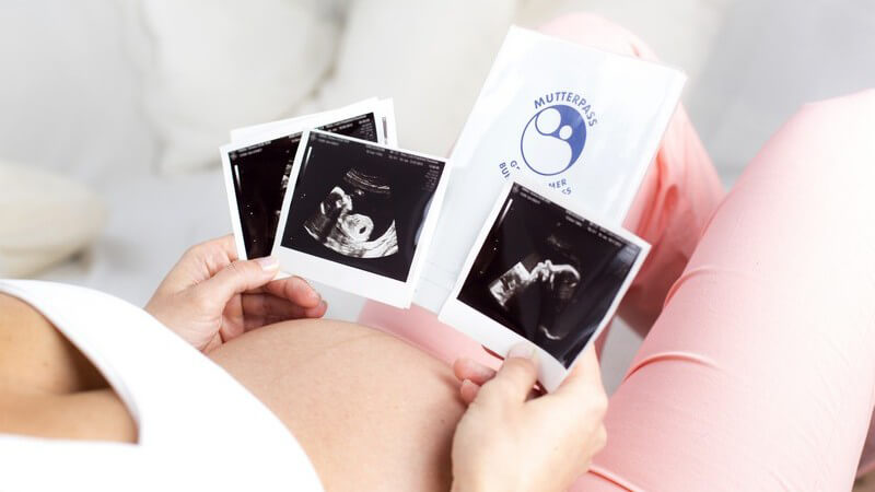Schwangere Frau in rosa Hose hält Mutterpass und Ultraschallbilder in den Händen