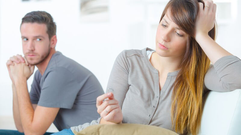 Junges Paar auf einer Couch, sie sitzt von ihm abgewendet und starrt bedrückt auf einen Ring in ihrer Hand