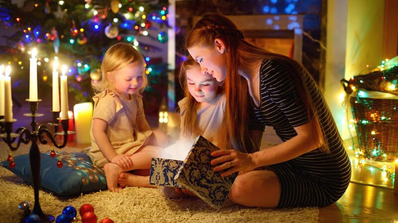 Junge Mutter sitzt mit ihren beiden kleinen Töchtern an Weihnachten auf dem Boden und packt Geschenke aus