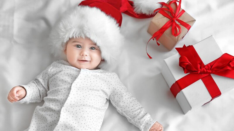 Baby in gepunktetem Schlafanzug liegt mit Weihnachtsmütze auf einer weißen Decke neben Geschenken mit roter Schleife