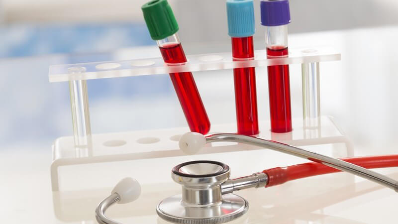 Drei Reagenzgläser mit Blutproben auf einem Ständer im Labor, davor liegt ein Stethoskop