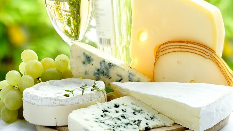 Verschiedene Sorten an Käse, daneben Trauben und Glas Weißwein