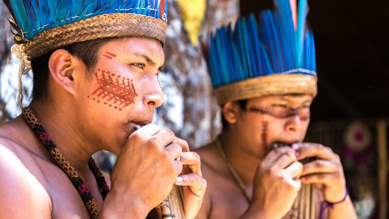 Zwei brasilianische Ureinwohner mit blauem Federschmuck spielen auf einer Holzflöte