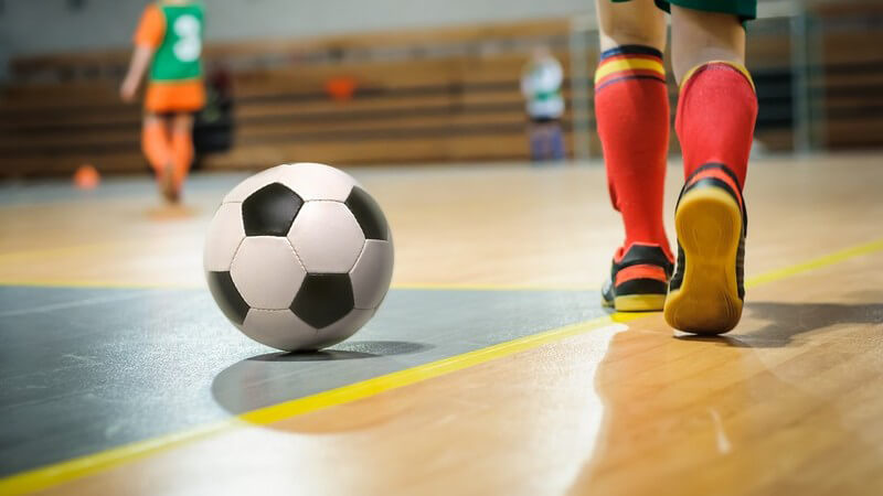 Ein Fußball liegt auf dem Hallenboden, daneben geht ein Junge in Fußballkleidung